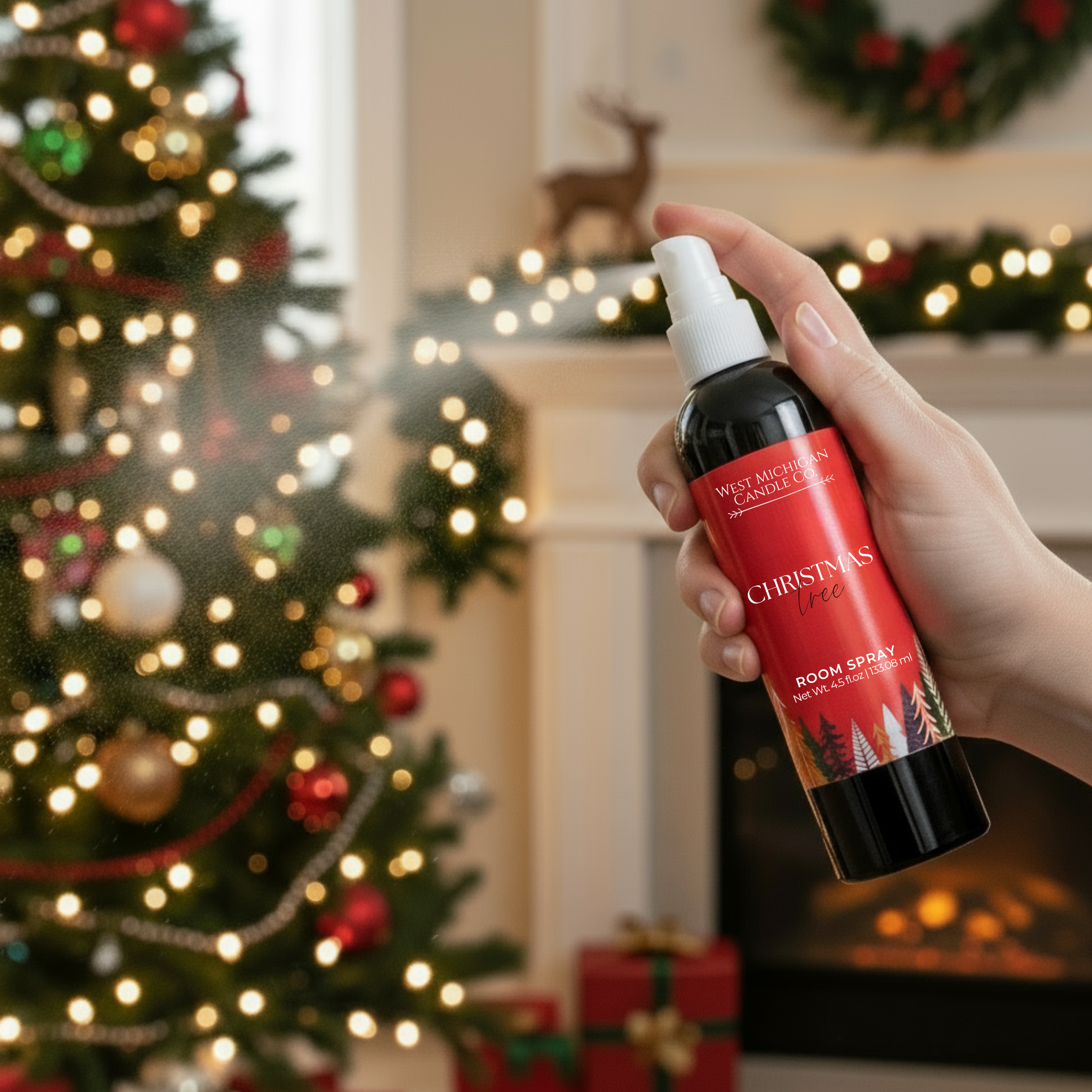 Hand holding a Christmas-themed room spray bottle in front of a decorated Christmas tree and fireplace.